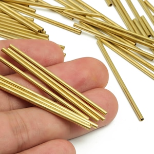 May include: A collection of small, gold-coloured metal tubes. Some are held in a hand, others are scattered in the background. The tubes are hollow and cylindrical, likely for jewellery making or crafting. The tubes are approximately 2.5 cm in length.
