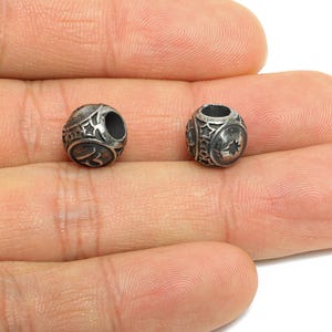 Peut inclure: Deux petites perles argentées avec des motifs complexes d'étoiles et géométriques. Chaque perle a un trou central et un motif détaillé, idéal pour la fabrication de bijoux ou les projets d'artisanat.