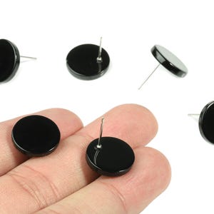 May include: Several black, round stud earrings with silver posts are displayed against a white background. One pair is held in a hand. The earrings have a glossy finish and a simple, minimalist design.