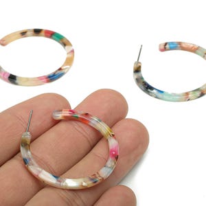 Pode incluir: Três brincos de argola coloridos com fecho de pino. Os brincos são feitos de um material acrílico marmorizado com tons de rosa, azul, verde e bege. Um brinco é segurado em uma mão.