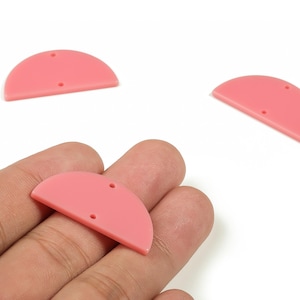 以下が含まれることがあります： ピンク色の半円形アクリルチャーム3つ。それぞれに2つの穴があります。1つは手に持たれ、他の2つは白い背景に散らばっています。ジュエリー作りやクラフトプロジェクトに最適です。