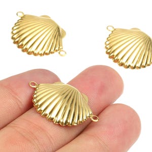 Puede incluir: Tres dijes dorados con forma de concha marina, con una textura acanalada y pequeños bucles para sujetarlos a la joyería. Un dije se sostiene en una mano, mientras que los otros dos se muestran arriba. Los dijes son de metal brillante.
