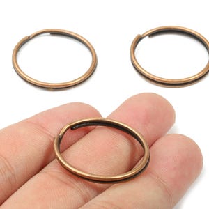 Puede incluir: Tres anillos partidos de color bronce, dos están colocados arriba y uno se sostiene en una mano. Los anillos son circulares con una ranura para sujetarlos a otros objetos. Los anillos tienen un interior oscuro y un exterior de bronce.