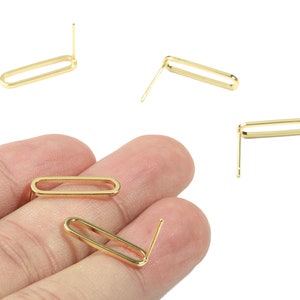 Puede incluir: Cinco hallazgos de pendientes de metal dorado con un bucle rectangular y un poste recto. Los hallazgos se muestran en una mano y sobre un fondo blanco.