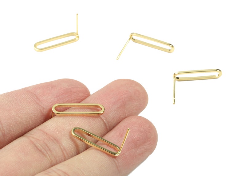 Puede incluir: Cinco hallazgos de pendientes de metal dorado con un bucle rectangular y un poste recto. Los hallazgos se muestran en una mano y sobre un fondo blanco.