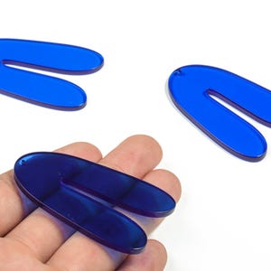 Op de afbeelding: Drie levendige blauwe, U-vormige acrylbedels. Elke bedel heeft een klein gaatje aan de bovenkant om te rijgen. Eén bedel wordt in een hand gehouden, terwijl de andere twee afzonderlijk tegen een witte achtergrond worden weergegeven. Deze bedels zijn ideaal voor het maken van sieraden.