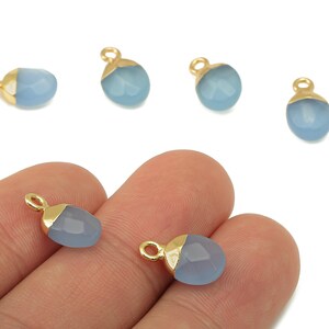 May include: Several light blue teardrop-shaped gemstone pendants with gold-colored caps and loops. Some pendants are scattered, while two are held in a hand. The gemstones have a faceted surface.