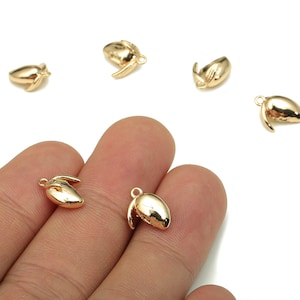 May include: Several small, gold-colored charms shaped like a seed or nut, with a loop for attaching to jewelry. The charms are scattered on a white background, with some resting on a person's fingers.
