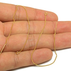 May include: A pair of gold-colored threader earrings. Each earring features a delicate chain with a small bar at one end and a loop at the other. The earrings are displayed against a hand.