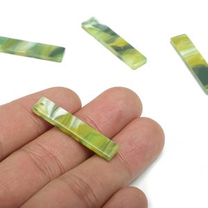 May include: Four rectangular beads with a hole at one end, in shades of green, yellow, and white. One bead is held in a hand. The beads have a marbled appearance and are likely for jewelry making or crafts.