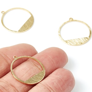 Pode incluir: Pingentes de metal dourado. Cada pingente apresenta um formato circular com uma seção diagonal texturizada. Os pingentes têm um pequeno laço na parte superior para prender em joias. A imagem mostra três pingentes, um deles na mão.