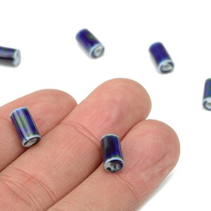 Könnte beinhalten: Sechs zylindrische blaue und weiße Perlen. Die Perlen sind dunkelblau mit weißen und grünen Akzenten. Die Perlen sind klein und scheinen aus Glas oder Keramik zu sein. Die Perlen sind auf einem weißen Hintergrund verstreut.
