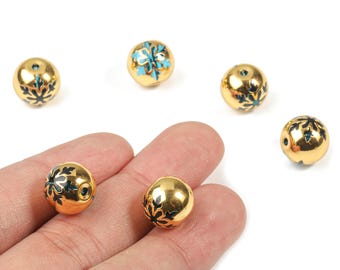 May include: Gold-colored round beads with black and teal floral designs. Several beads are scattered on a white surface, with two held in a hand. These decorative beads are suitable for jewelry making.