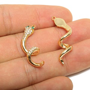 May include: Two gold-coloured snake charms. One charm features a head with clear and green gemstones, while the other is a plain gold snake. The charms are curled and have a loop at the top for hanging.