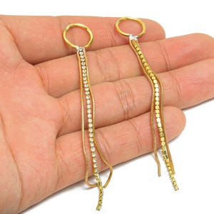 May include: A pair of gold-toned dangle earrings. Each earring features a circular top, a section with clear crystals, and a chain-like design. The earrings are displayed against a neutral background.