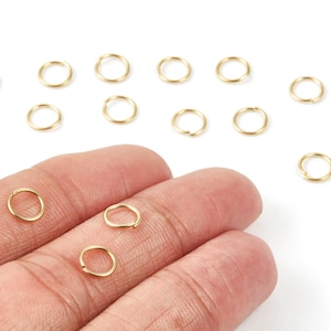 May include: A collection of small, gold-colored jump rings, some scattered and others held in a hand. These jewelry-making components are circular with a small opening, ideal for connecting other jewelry pieces. The rings are displayed against a white background.