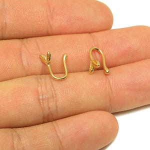 May include: Two gold-colored nose cuffs, each with a leaf design. The cuffs are in a U-shape, with a small leaf detail on one end and a rounded tip on the other. The jewelry is displayed on a light skin tone.