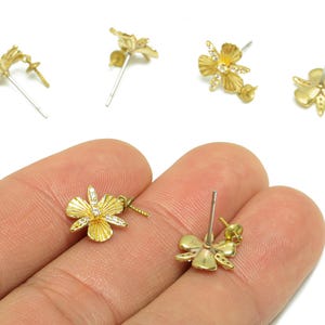 Pode incluir: Brincos de botão dourados em forma de flor com fecho de pino. Os brincos têm uma superfície texturizada e são adornados com pequenos cristais transparentes. Vários pares de brincos são visíveis.