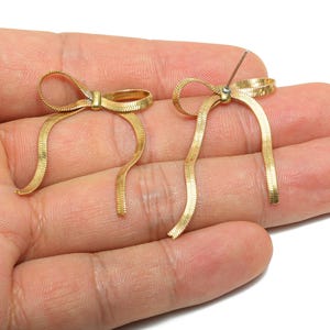 May include: A pair of gold-colored bow earrings. Each earring features a bow shape with long, dangling ribbon-like strands. The earrings are made of a textured, woven material and are displayed against a hand.