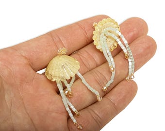 May include: A pair of handmade earrings with a jellyfish design. The earrings feature a translucent yellow dome, white beaded tentacles, and gold-colored accents. The earrings are held in a hand against a white background.
