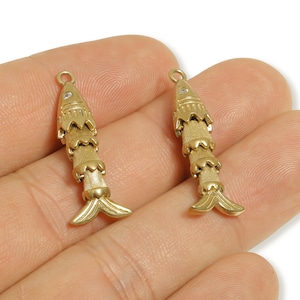 May include: Two gold-colored fish-shaped charms. Each charm has a textured body, a small gem, and a loop at the top for hanging. The fish have detailed fins and tails, suitable for jewelry making or crafting.