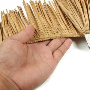 May include: Close-up of a decorative fringe trim in a natural tan color. The fringe is made of thin, textured strips attached to a woven base. The trim is held by a hand against a white background, showcasing the texture and detail.