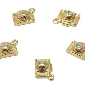 May include: Several gold-colored camera charms with a clear, round gem in the center of the lens. Each charm has a small loop at the top for attaching to a chain or bracelet. The charms are on a white background.
