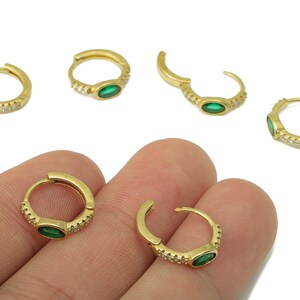 Op de afbeelding: Goudkleurige oorringen met een centrale groene edelsteen en kleine heldere stenen. De oorbellen worden in verschillende posities getoond, sommige open en sommige gesloten. De oorbellen worden in een hand gehouden.