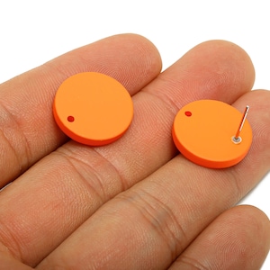 Pode incluir: Dois brincos redondos laranja. Um brinco tem um pino e uma parte traseira, o outro tem um buraco para o pino. Os brincos são segurados na mão de uma pessoa contra um fundo branco. Os brincos são de cor laranja brilhante.