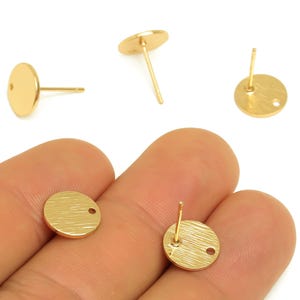 Può includere: Orecchini a bottone rotondi color oro con superficie strutturata. Gli orecchini hanno un perno e un piccolo foro per il fissaggio. Diversi orecchini sono esposti su sfondo bianco.