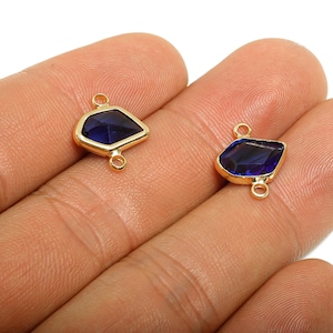 Pode incluir: Dois conectores de pedras preciosas douradas com uma pedra azul escura de formato irregular. Cada conector tem uma argola na parte superior e inferior para fixar a outros componentes de joalheria. As pedras preciosas são presas em uma moldura dourada.