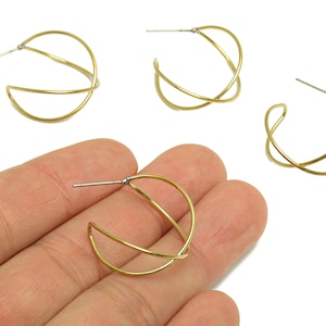 Op de afbeelding: Goudkleurige oorringen met een uniek, kruisend ontwerp. De oorbellen hebben een stift sluiting en worden getoond in een groep van vier, waarvan er één in de hand wordt gehouden. De oorbellen zijn gemaakt van dun, gebogen metaal.