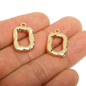 May include: Two gold-colored, open-frame rectangle charms with a textured surface. Each charm has a small loop at the top for attaching to a chain or other jewelry component. The charms are displayed against a neutral background.