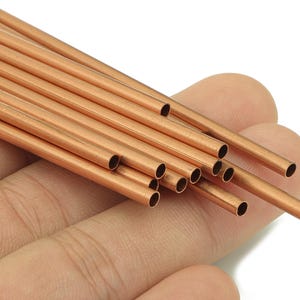May include: A selection of copper tubes, each with a hollow centre. The tubes are a warm, reddish-brown colour and are held in a hand. These small tubes are likely used for jewellery making or other craft projects.