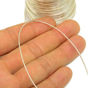 May include: A spool of silver wire, with a strand of wire held in a hand. The wire is thin and appears to be made of a metal. The background is white.
