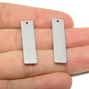 Puede incluir: Dos colgantes rectangulares de metal plateado, con un pequeño agujero en la parte superior. Los colgantes se sostienen en una mano sobre un fondo blanco. Adecuado para la fabricación de joyas.