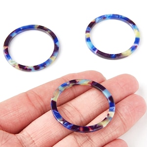 May include: Three colorful, round acrylic jewelry components. The rings feature a marbled pattern of blue, purple, and yellow. The components are shown against a white background. The jewelry components are held in a hand.