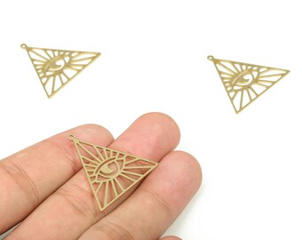 Peut inclure: Trois breloques triangulaires dorées avec un motif d'œil. Les breloques sont découpées avec un motif de rayons de soleil autour de l'œil. Une breloque est tenue dans une main. Les breloques ont une petite boucle en haut pour la joaillerie.