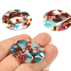 May include: Three oval-shaped beads with a marbled design. The beads feature a mix of colors, including teal, brown, white, and gold flecks. One bead is held in a hand, showcasing the pattern and texture.