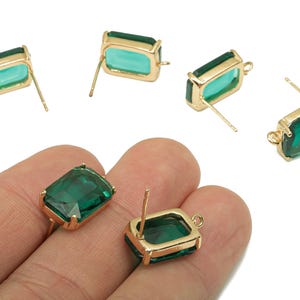 May include: Emerald-cut green gemstone stud earrings with gold-colored metal settings. The earrings have a rectangular shape and a post for pierced ears. Several earrings are visible, some held in a hand.