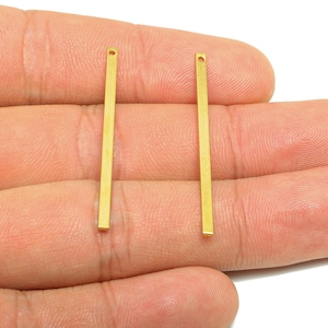 May include: Two gold-colored, rectangular bar charms with a small hole at the top. The charms are thin and straight, and are displayed against a light background.