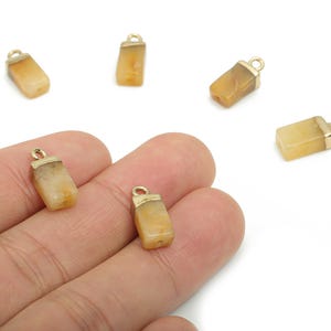 Puede incluir: Seis pequeños colgantes rectangulares de piedra preciosa amarillo-anaranjada con tapas y lazos de metal dorado. Los colgantes están dispersos sobre un fondo blanco, uno de ellos sostenido en la mano de una persona. Ideal para hacer joyas.