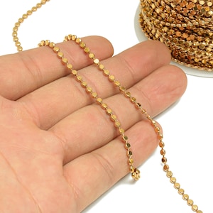 May include: A gold chain with small, round links. The chain is shown on a hand, and a spool of the chain is in the background.