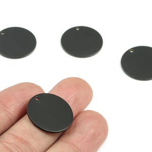 Puede incluir: Cuatro colgantes redondos de metal negro con un pequeño agujero en la parte superior para ensartar. Un colgante ovalado se sostiene en una mano. Los colgantes son de color negro mate sólido y se muestran sobre un fondo blanco.