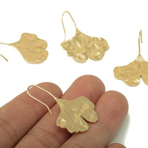 Puede incluir: Pendientes dorados con forma de hoja de ginkgo con superficies texturizadas. Los pendientes se muestran sobre un fondo blanco, con un pendiente en la mano. Los pendientes tienen un diseño de gancho.