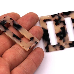 Puede incluir: Pendientes rectangulares con estampado de carey y centro abierto. Los pendientes son de color beige claro con manchas marrones oscuras y negras. Un pendiente se sostiene en una mano, y hay dos más al lado.