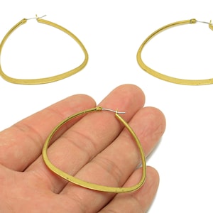 May include: Gold-toned, triangular hoop earrings. The earrings have a flat, slightly textured surface and a simple, minimalist design. The image shows three earrings, with one held in a hand. The earrings are a warm, metallic color.