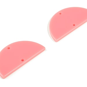 Może przedstawiać: Dwa różowe, półokrągłe wisiorki akrylowe z dwoma otworami każdy. Wisiorki mają jednolity, nieprzezroczysty kolor i gładkie, błyszczące wykończenie. Są pokazane na białym tle, idealne do tworzenia biżuterii lub rękodzieła.