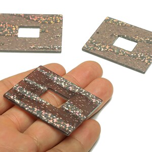 Op de afbeelding: Drie rechthoekige, bruine decoratieve kralen met een vierkante uitsparing in het midden. De kralen hebben een glitterig, gespikkeld ontwerp met roze, gouden en zilveren accenten. Eén kraal wordt in een hand gehouden.