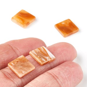 Könnte beinhalten: Vier quadratische, orange-braune Harzperlen mit Marmoreffekt. Jede Perle hat ein kleines Loch zum Auffädeln. Die Perlen werden vor weißem Hintergrund gezeigt.